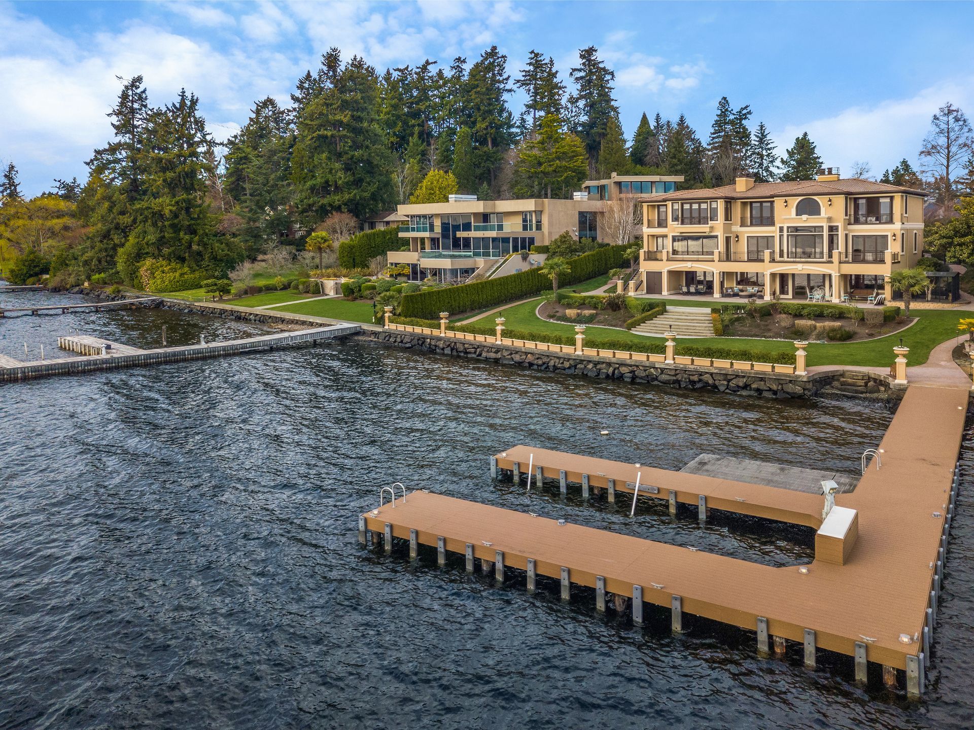 Docks extend into a dark body of water in front of luxury waterfront homes. Lush green trees line the hillside.
