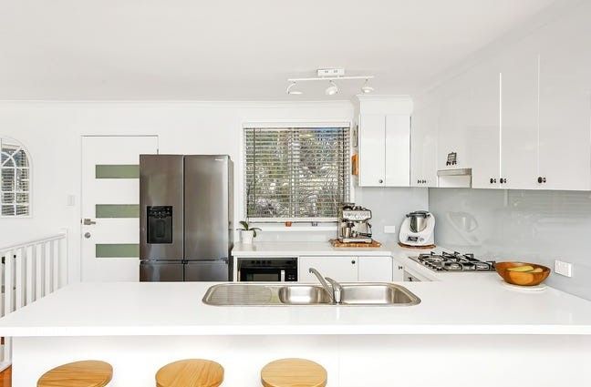 Modern, white kitchen with stainless steel appliances, countertops, and wooden stools at the island — Wards Kitchens in Berkeley Vale, NSW