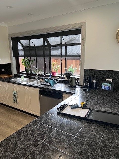 Kitchen with dark countertops, white cabinets, and a window with blinds; plants sit on the windowsill — Wards Kitchens in Berkeley Vale, NSW
