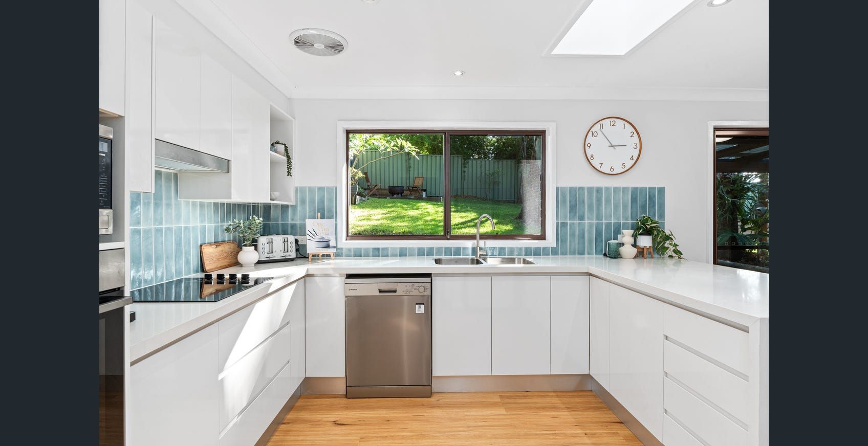 Bright white kitchen with blue tiled backsplash, stainless steel appliances, and a window overlooking a backyard — Wards Kitchens in Berkeley Vale, NSW