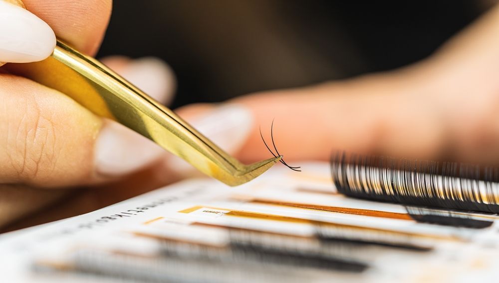 Eyelash extension application with tweezers and white under-eye pads on a client’s closed eye
