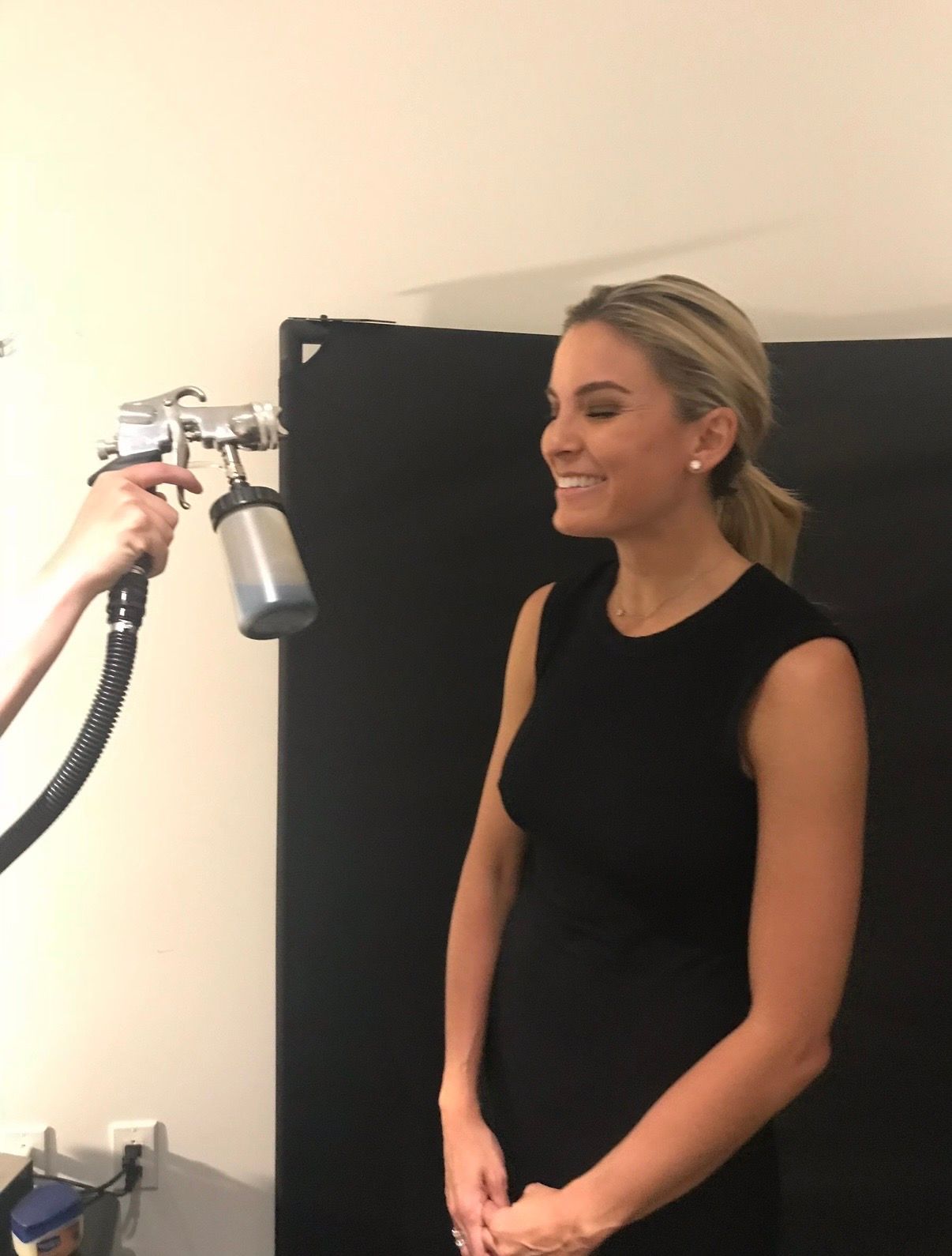 Woman in black sleeveless top posing for a portrait as a spray tan machine is aimed at her face
