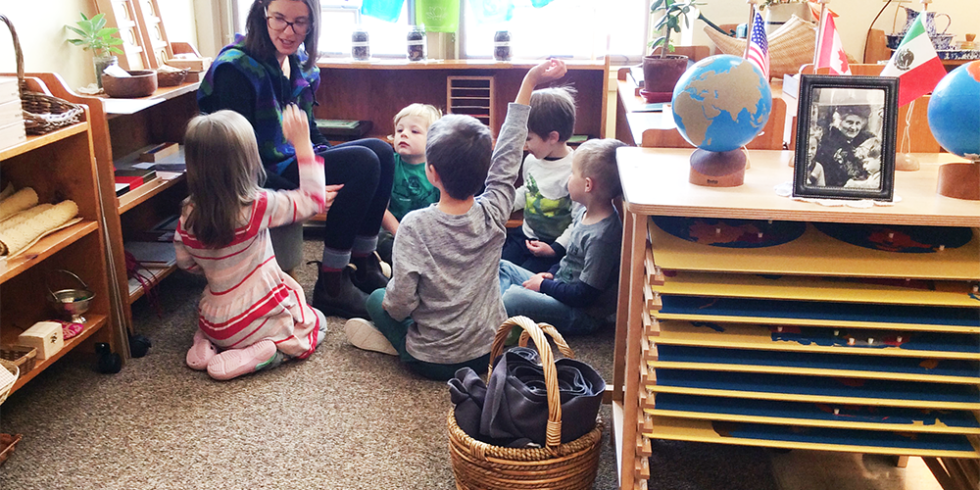 A group of children are sitting on the floor in a classroom with their hands up.