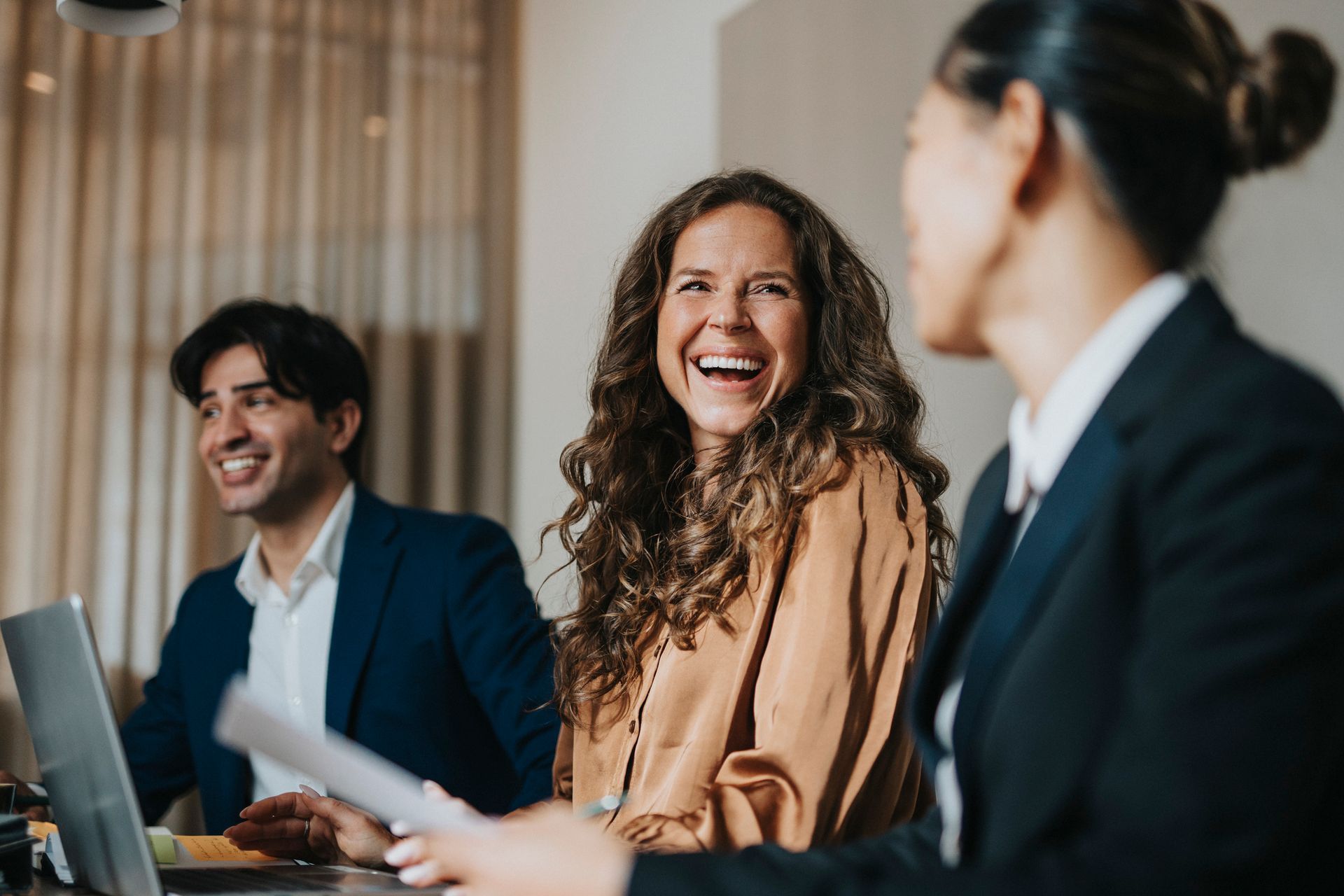 Three people laughing at a table, two men and one woman. They are in a modern office setting.