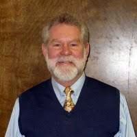 Man with a white beard and a navy vest, smiling, against a brown background. Man with a white beard and a navy vest, smiling, against a brown background.