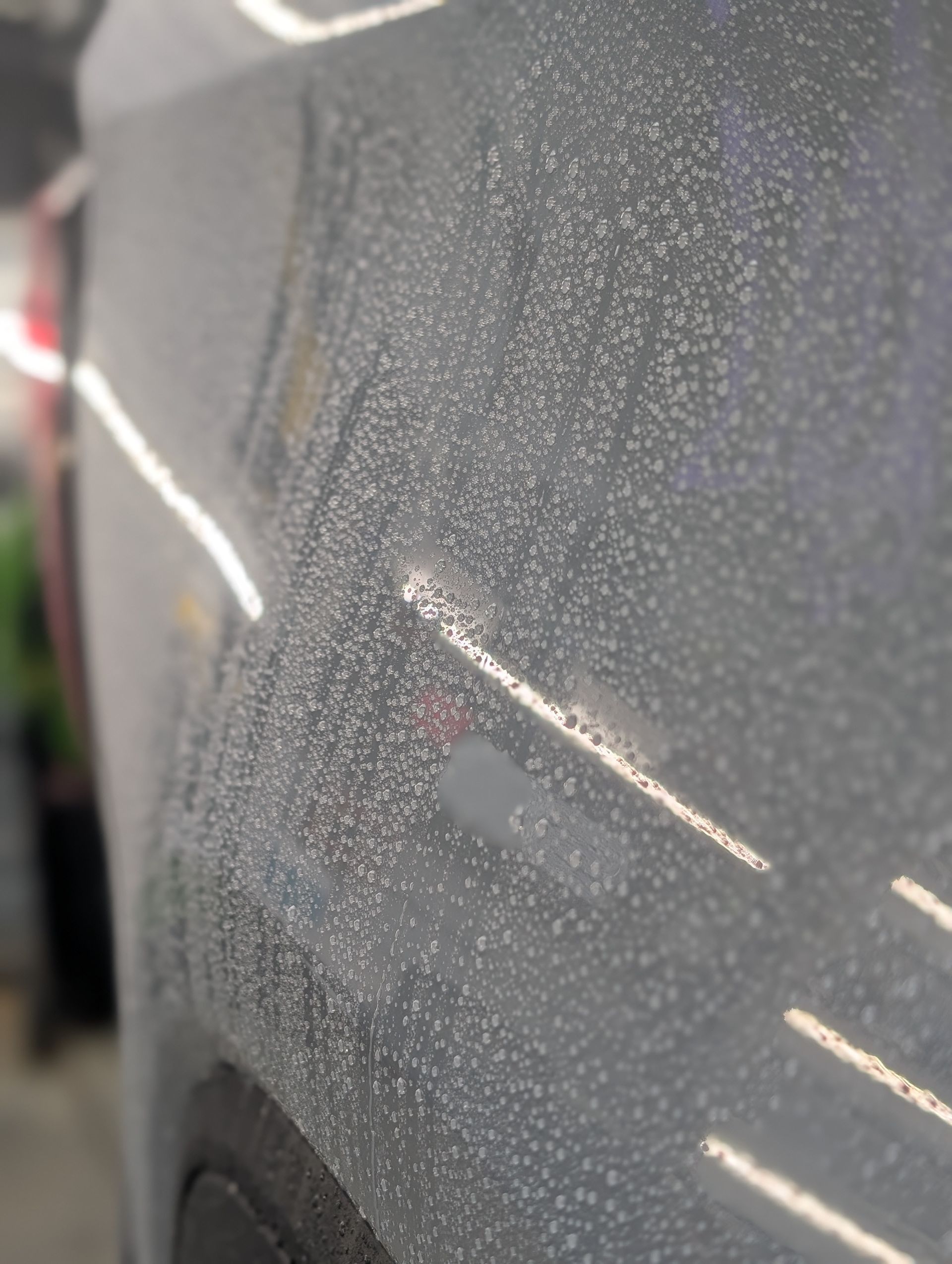 Close-up of a silver car door covered in small, water-like droplets, reflecting bright lights.