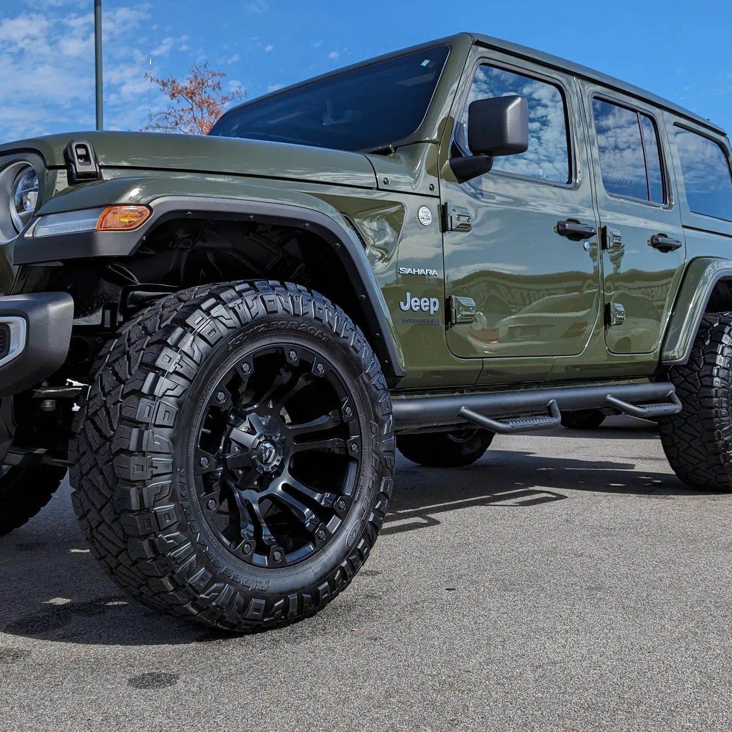 Green Jeep Wrangler with black wheels, parked on asphalt, under a blue sky.