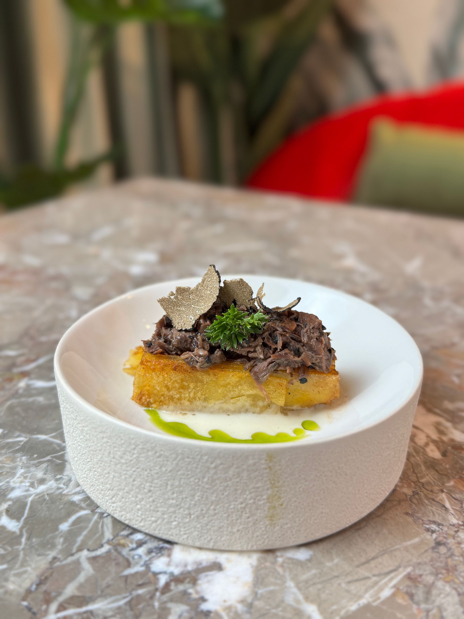 Una fetta di pane tostato croccante e dorata, guarnita con carne sfilacciata e scaglie di tartufo, servita su un piatto bianco con olio alle erbe aromatiche.