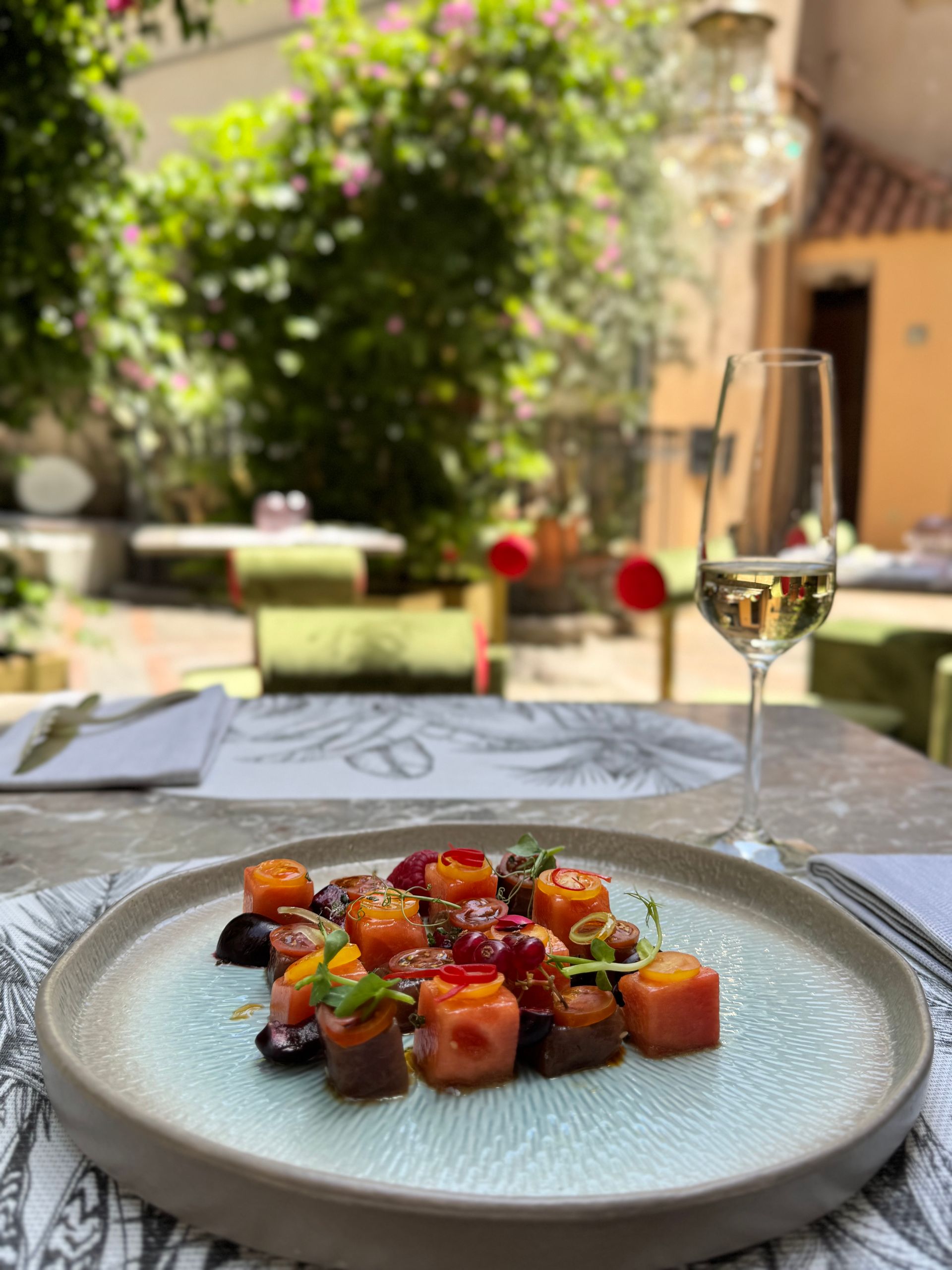 Un piatto gourmet a base di salmone a cubetti e insalata di barbabietole, servito su un piatto dalla consistenza particolare, accanto a un bicchiere di vino, in un cortile soleggiato.