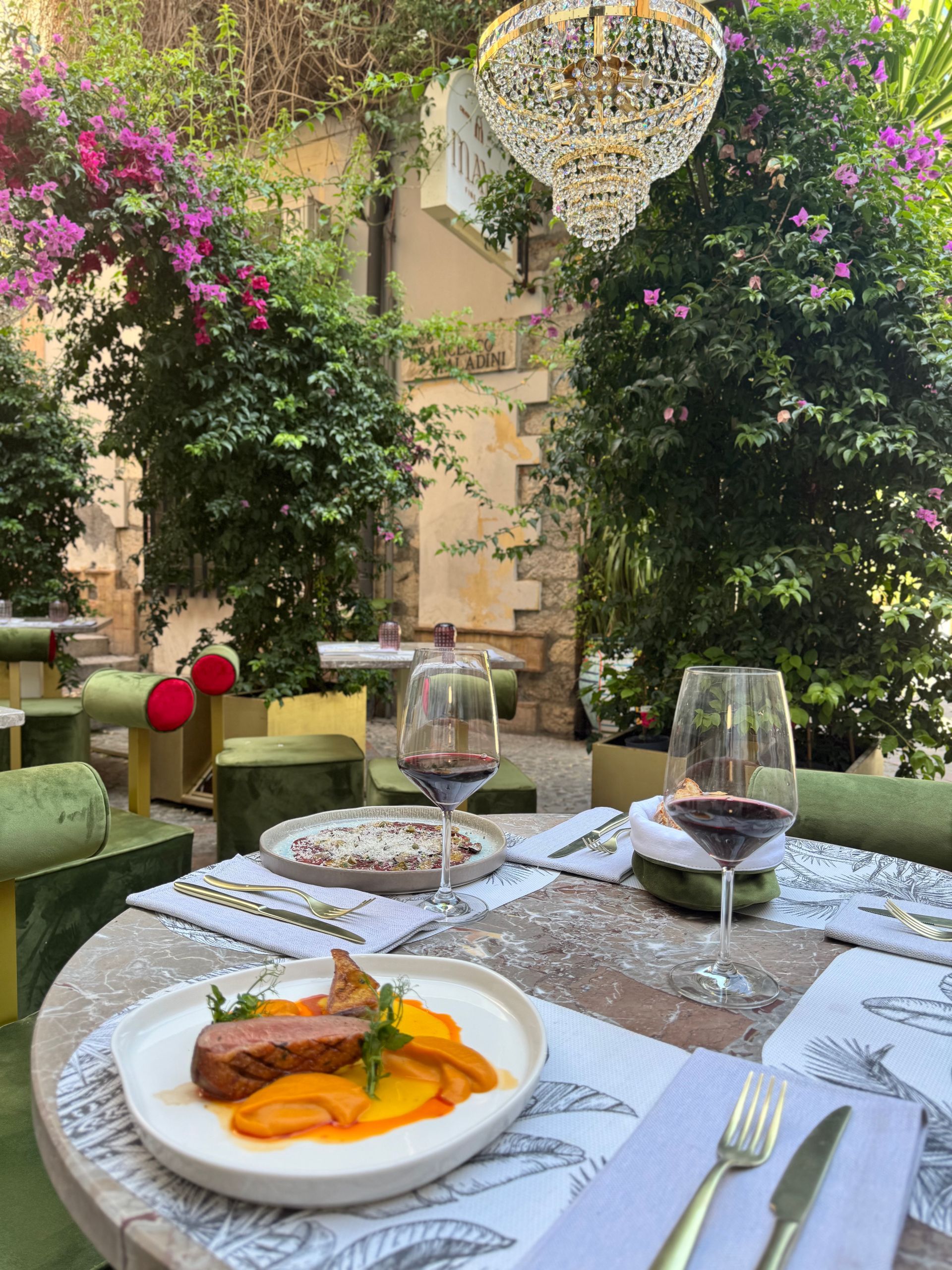 Un piatto di carne cotta e fette d'arancia è appoggiato su un tavolo con dei bicchieri da vino, all'aperto, in un ristorante immerso nel verde.