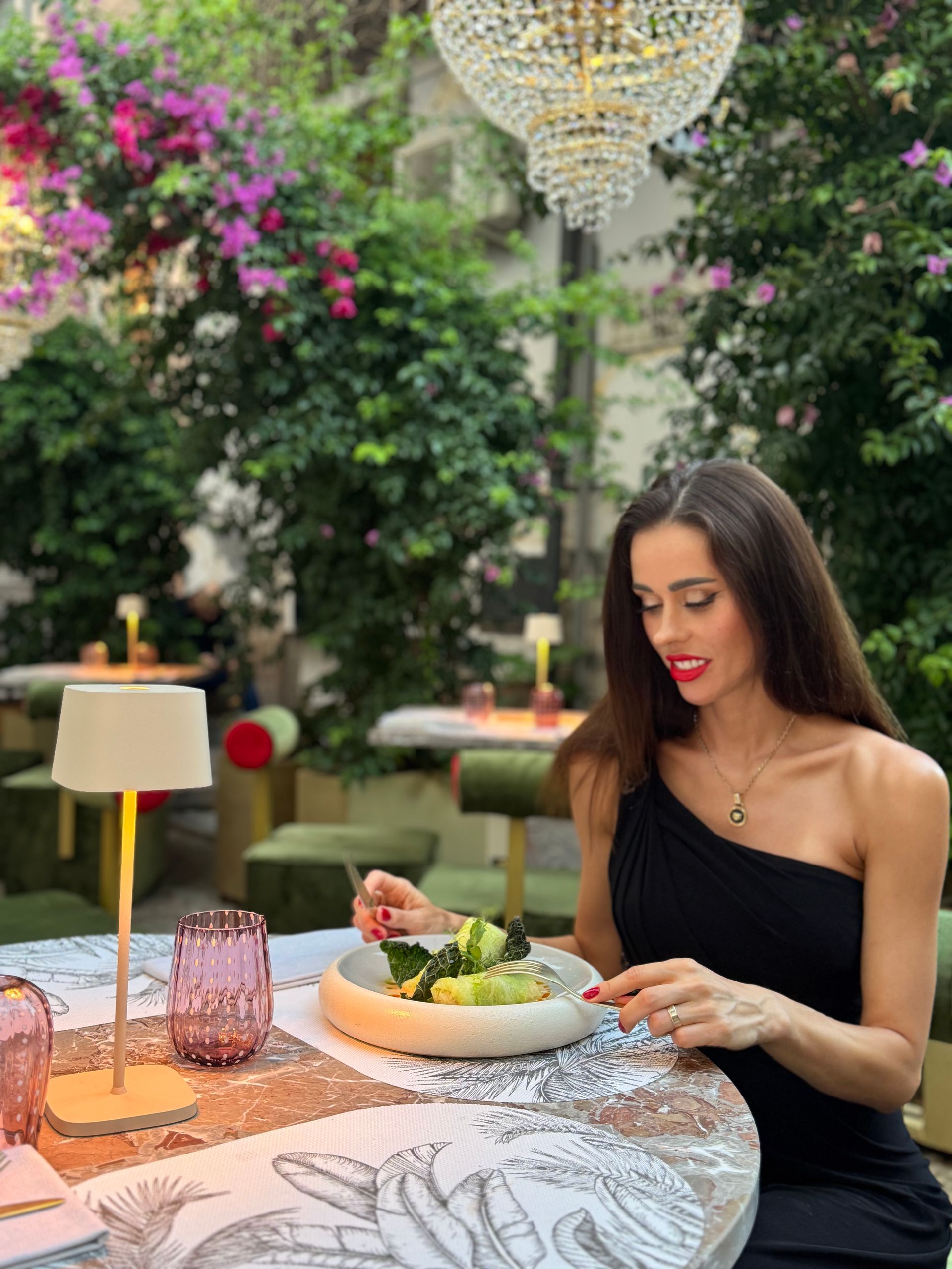 Una persona con indosso un abito nero monospalla mangia un'insalata a un tavolo all'aperto illuminato da candele, circondata da una vegetazione lussureggiante.