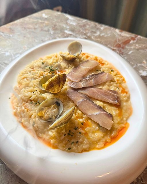 Un piatto bianco di cremoso risotto guarnito con tre piccole vongole e diverse fette di pesce affumicato, il tutto condito con un filo d'olio.
