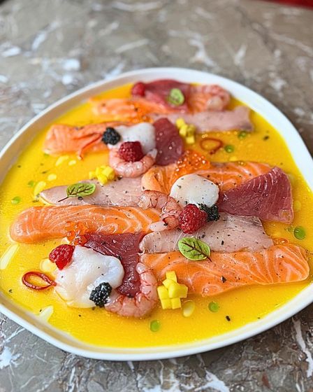 Un piatto vivace con un assortimento di fette di pesce crudo, gamberi, lamponi e cubetti di mango, servito in una salsa agli agrumi di un giallo brillante.