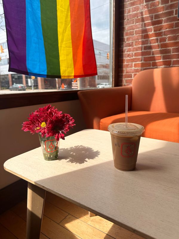 Rainbow flag in a cafe window above a table with flowers and iced coffee