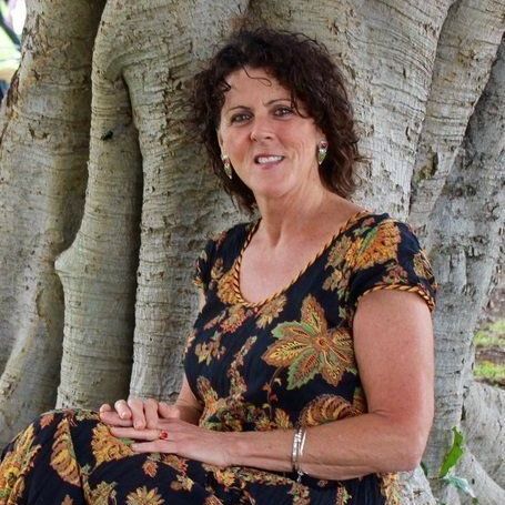 Woman leaning against a tree trunk, wearing a black floral top, smiling outdoors.