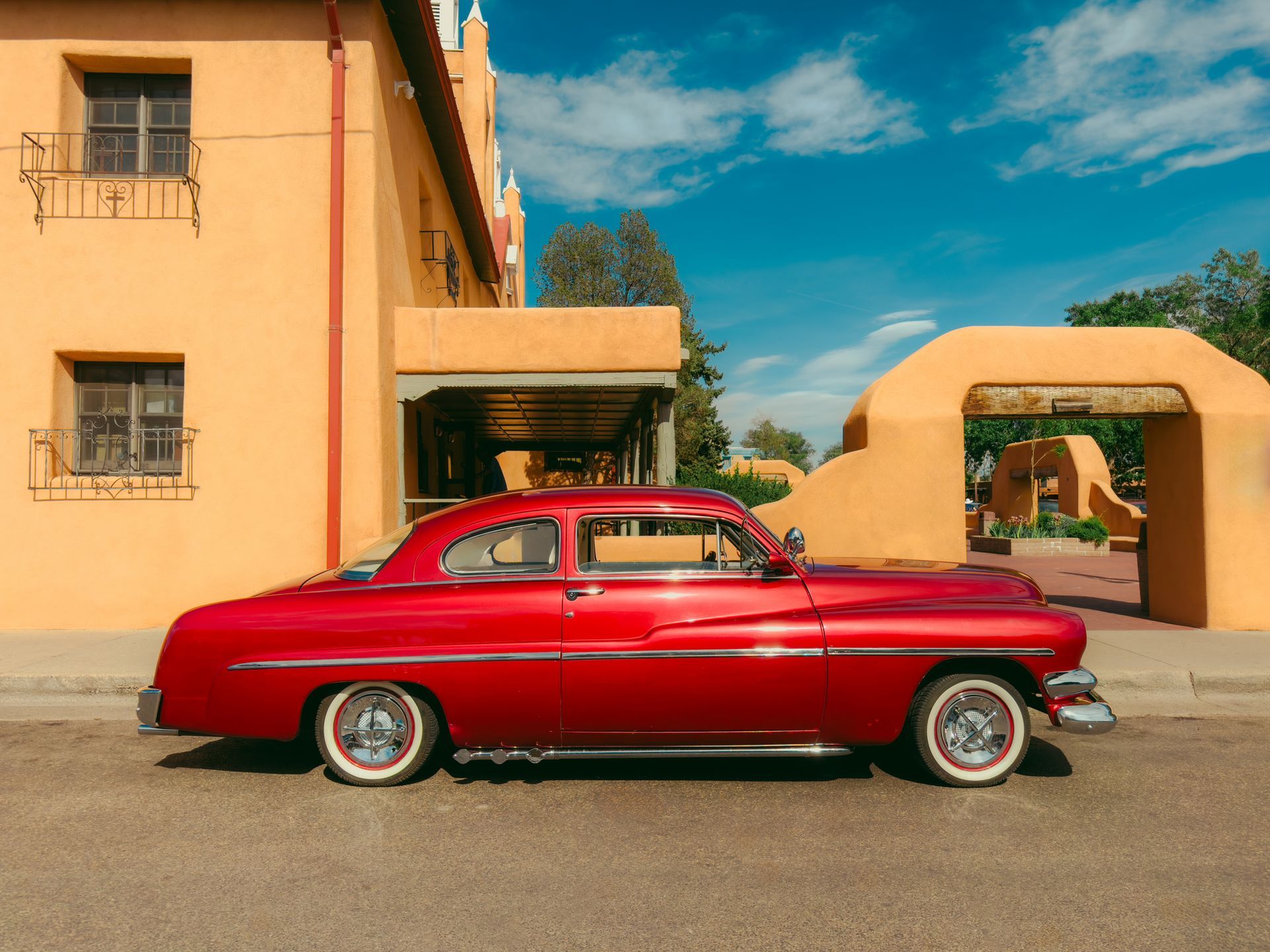 Red classic car parked in front of a tan building with an archway under a blue sky.