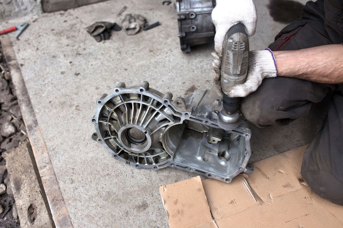 Mechanic using a power tool to work on a disassembled metal car part outdoors on concrete.