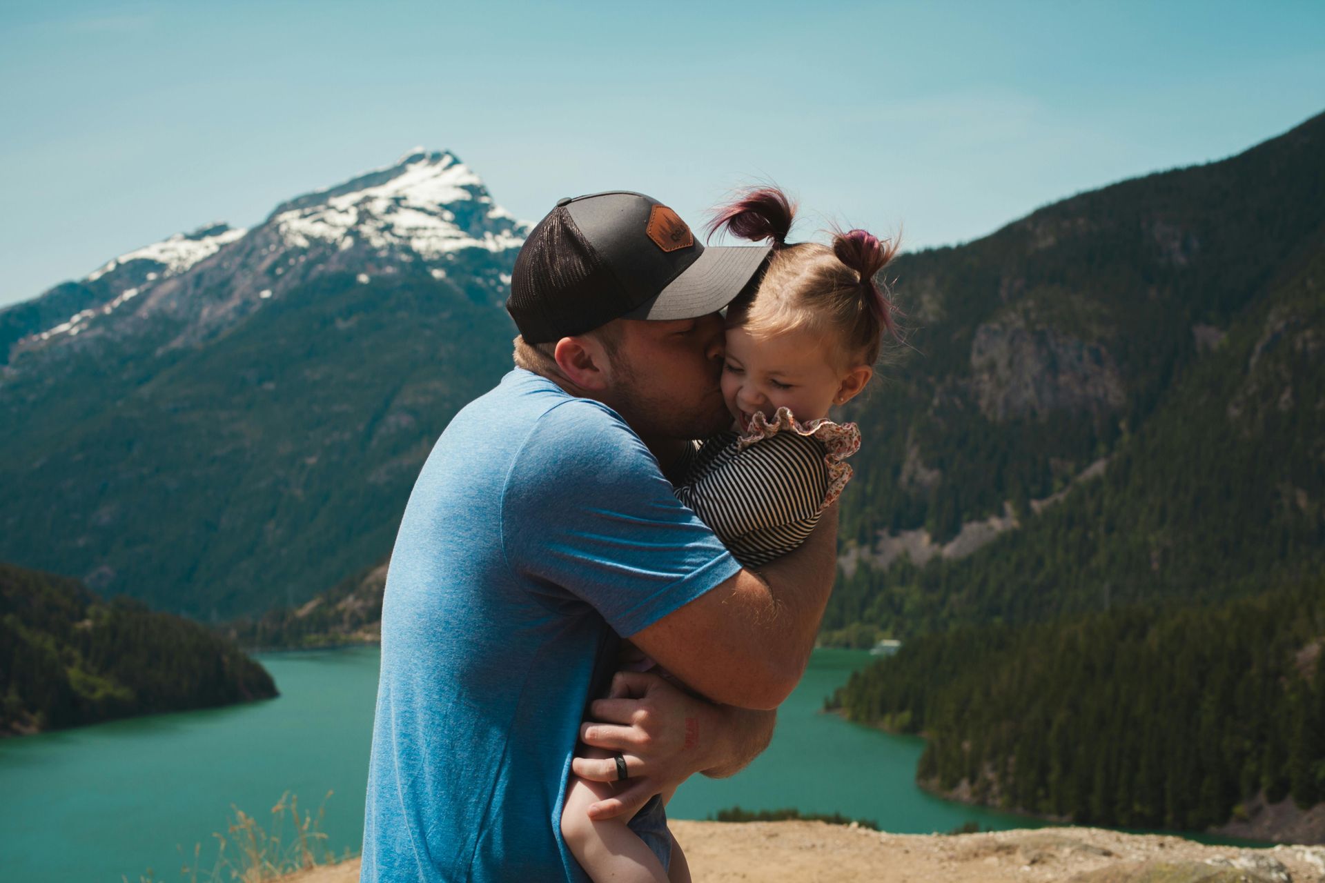 Man kisses baby on the cheek, mountains and lake in background.