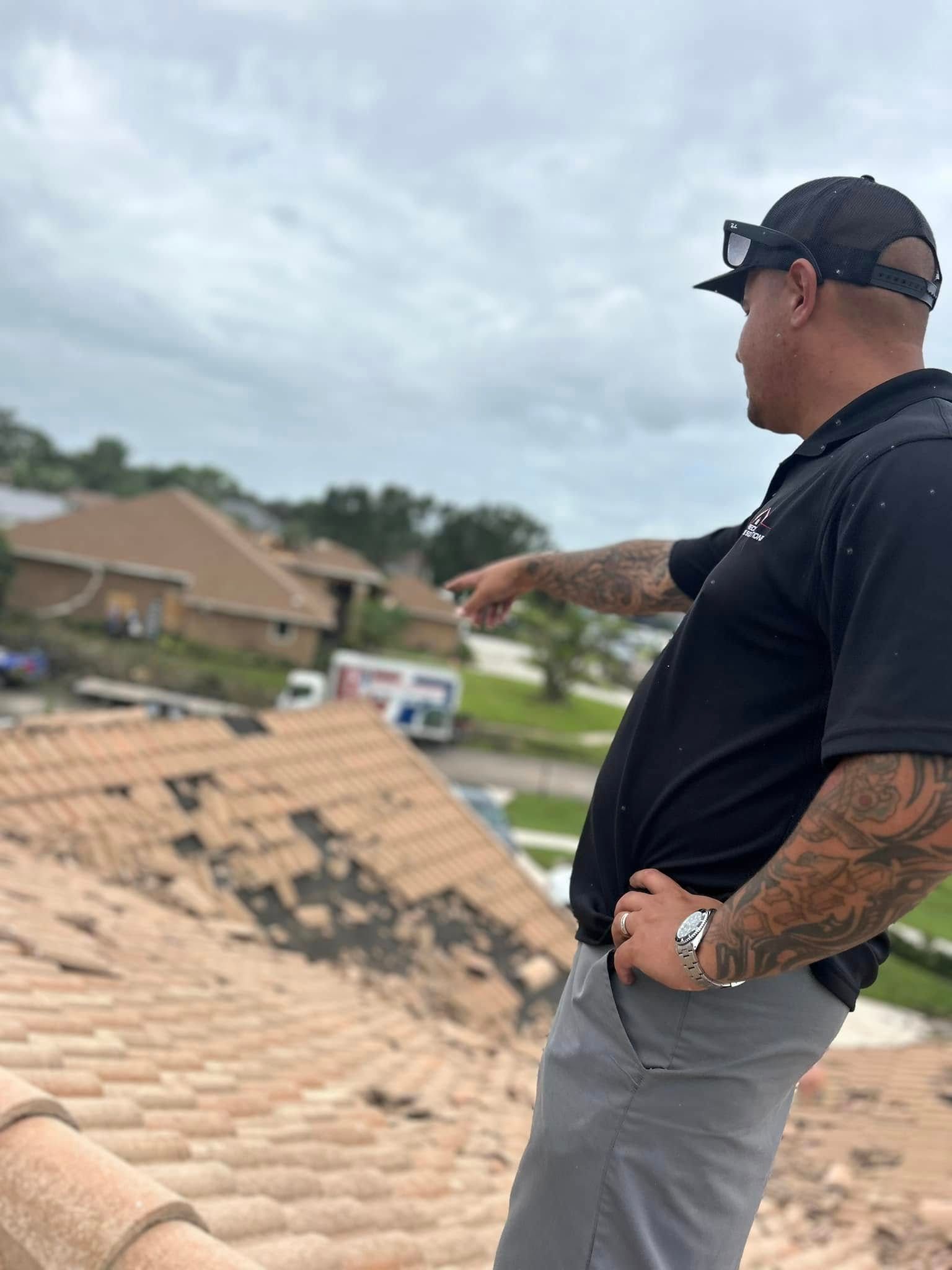 A person in a black polo shirt and cap stands on a tile roof, pointing toward a residential area with missing shingles.