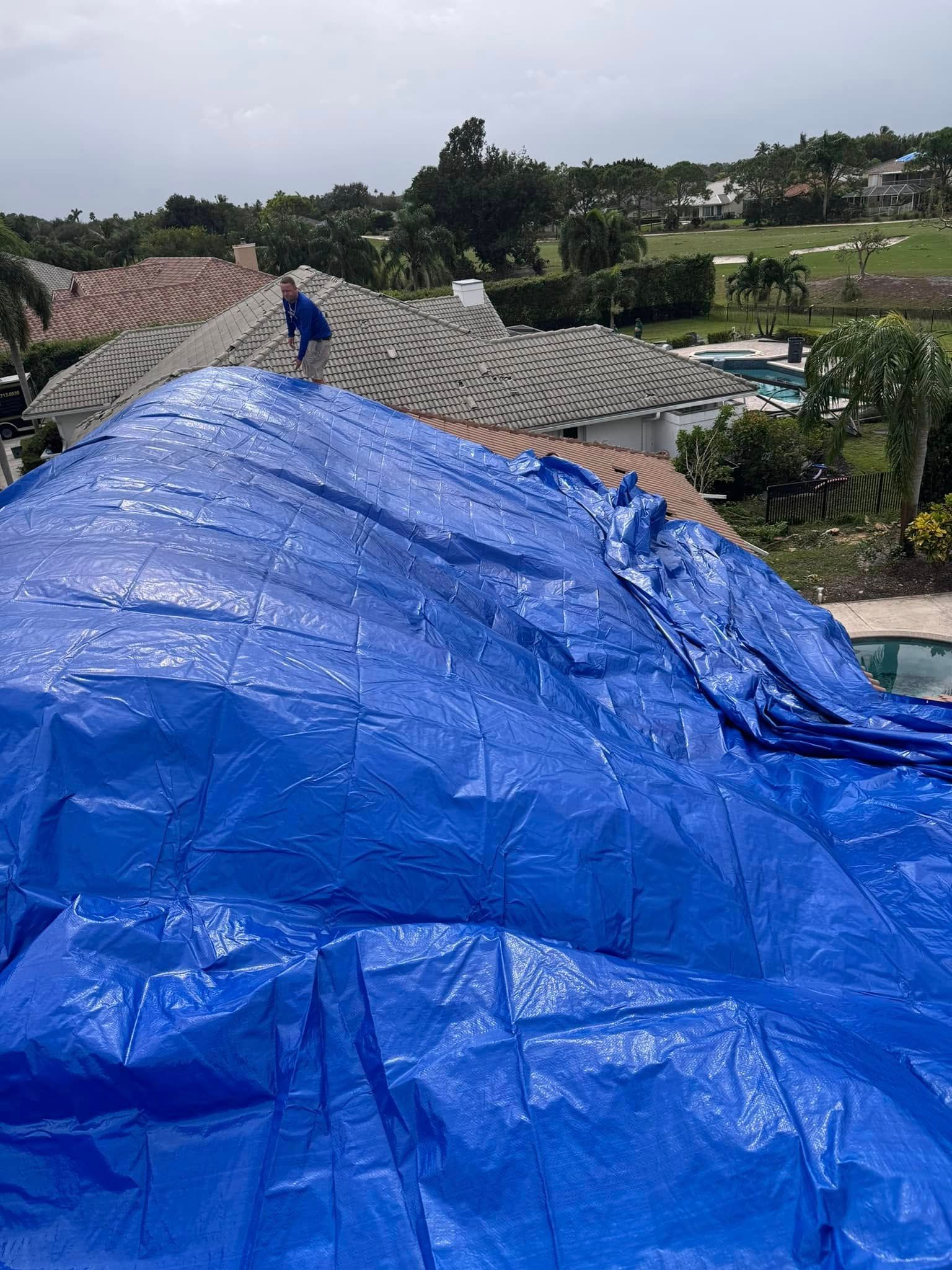 A person standing on a residential tile roof partially covered by a large blue tarp, overlooking a green yard and pool.