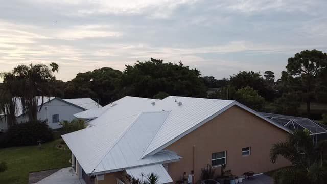 An aerial view of a suburban house with a bright white metal roof and tan exterior walls, surrounded by trees.