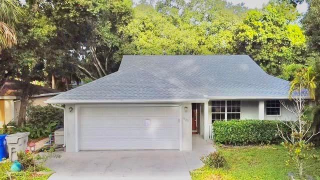 A one-story suburban home with a white garage, a light-colored exterior, a grey shingled roof, and green landscaping.