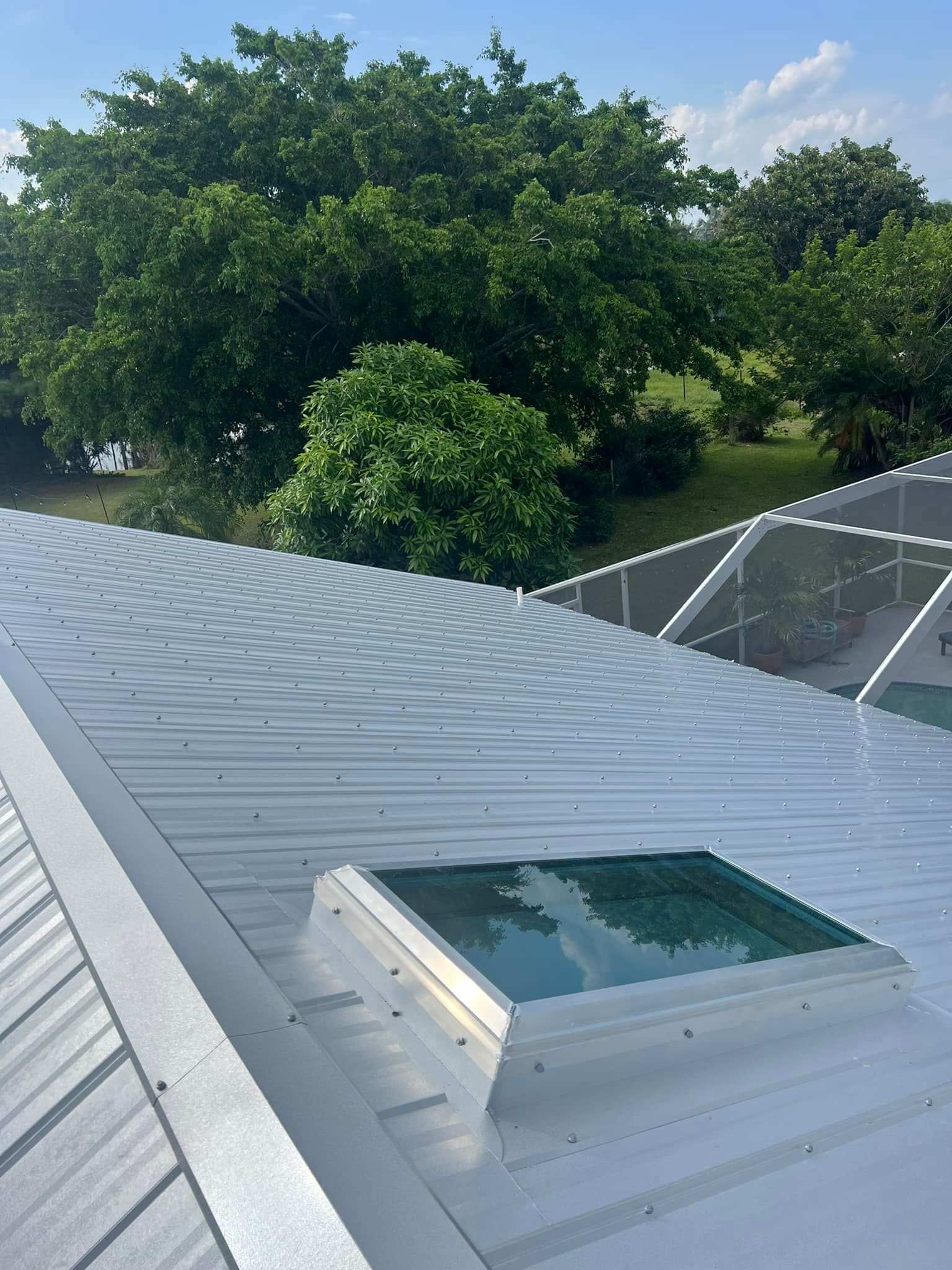 A close-up of a silver metal roof with a skylight, with lush green trees and a pool enclosure visible in the background.
