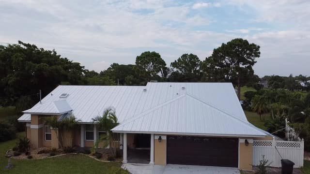 An aerial view of a single-story house with a light-colored metal roof, tan siding, and a dark two-car garage.