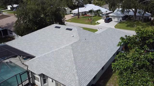 An aerial view of a gray shingled roof on a suburban house, with a screened-in pool and neighboring homes nearby.