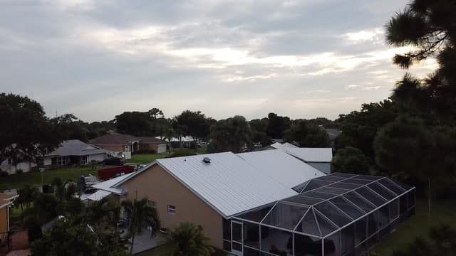 An aerial view of a tan house with a white roof and screened-in patio, surrounded by trees under a cloudy sky.