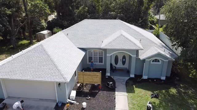 An aerial view of a light blue, single-story suburban house with a gray shingled roof, a front walkway, and a garage.