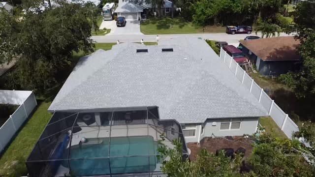 An aerial view of a suburban house with a gray shingled roof, a screened-in pool, and a white fence in a residential area.