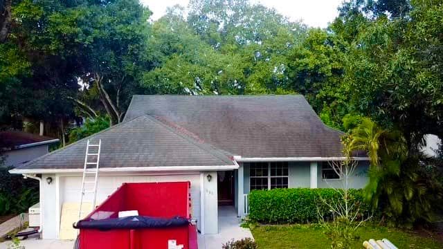 A house with a shingled roof under repair, featuring a large red dumpster in the driveway and a ladder leaning against it.