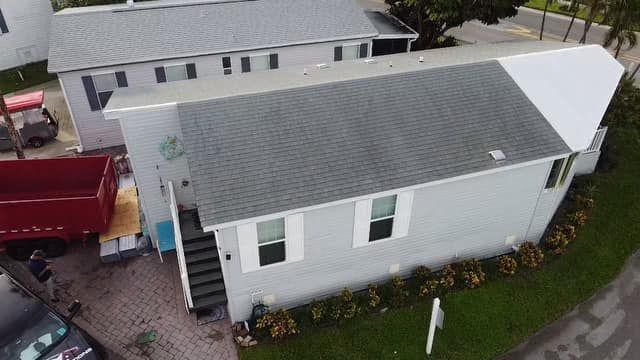 An elevated view of a gray residential house with a new asphalt roof next to a red dumpster and a paved driveway.