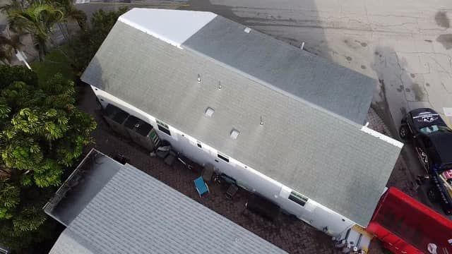 An aerial view of a rectangular house with a grey shingled roof, alongside a smaller structure and a red trailer.