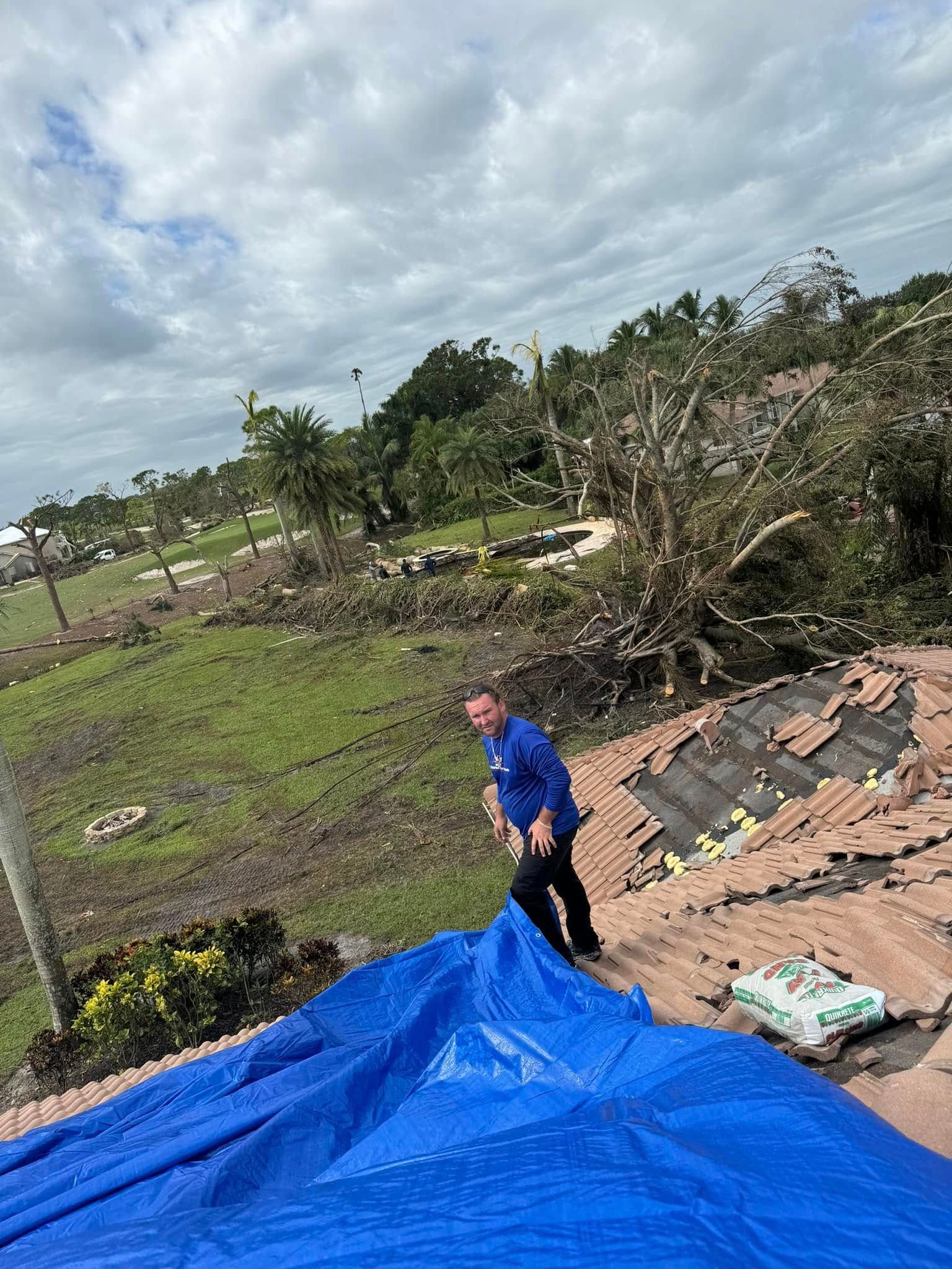 A person in a blue shirt secures a blue tarp over a damaged tile roof following a storm, with debris in the yard behind.
