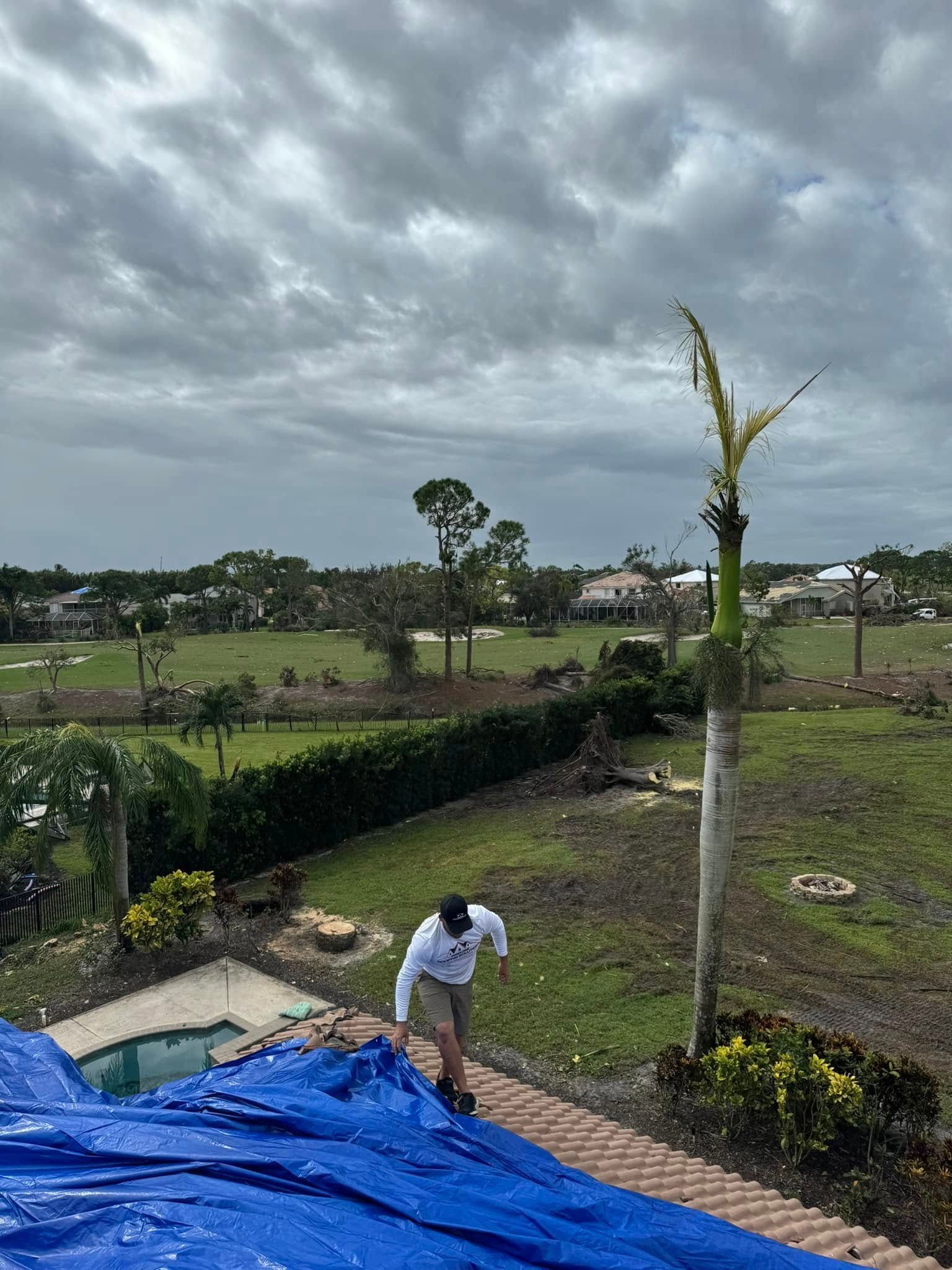 A person stands on a brick patio next to a blue tarp-covered pool area, overlooking a landscape with storm-damaged trees.