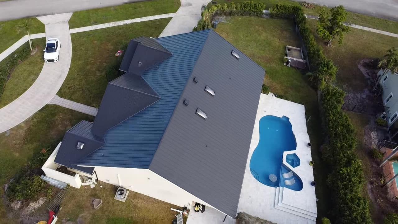 Aerial view of a residential house with a dark metal roof, a backyard pool, and a white car in the driveway.