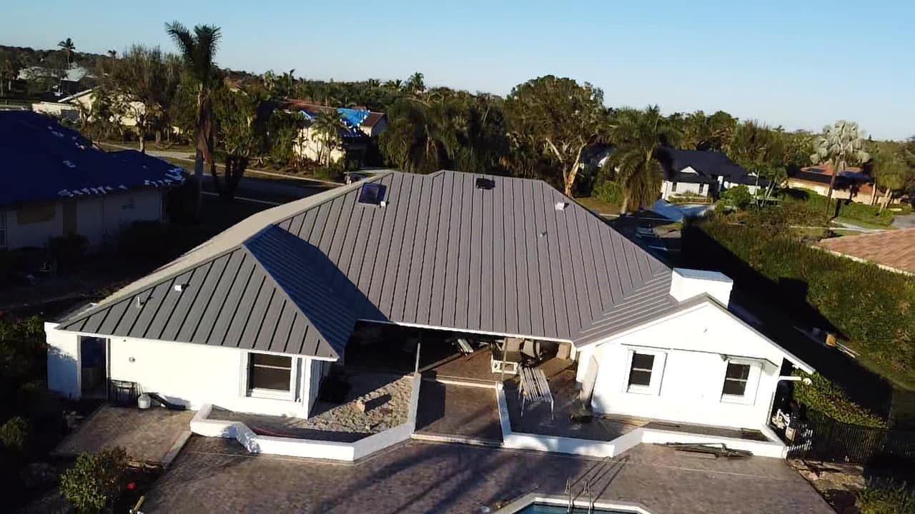 An aerial view of a white, single-story residential home with a large, grey, textured metal roof and a backyard pool.