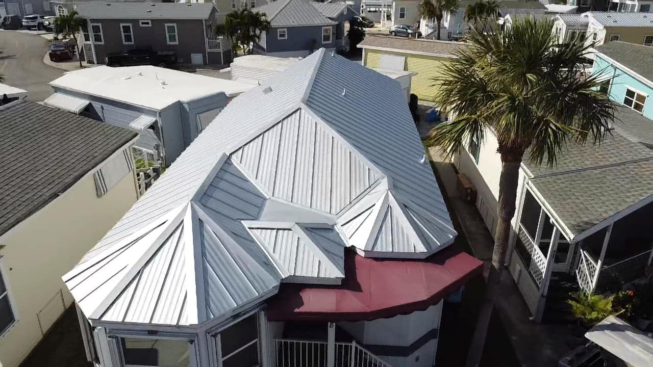 Aerial view of a coastal home with a shiny, light-gray metal roof and a maroon awning, surrounded by neighboring homes.