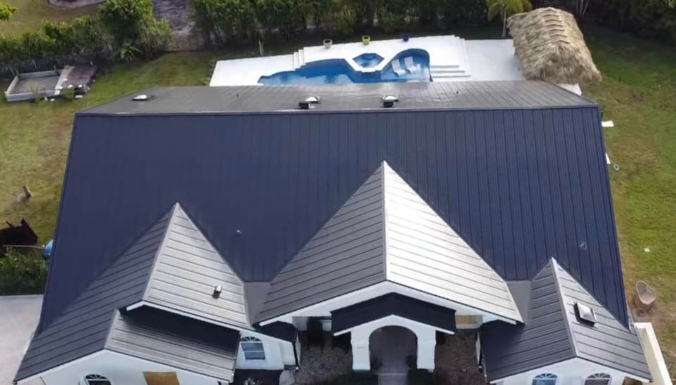 An aerial view of a house with a dark metal roof, featuring multiple gables and an adjacent pool area with a gazebo.