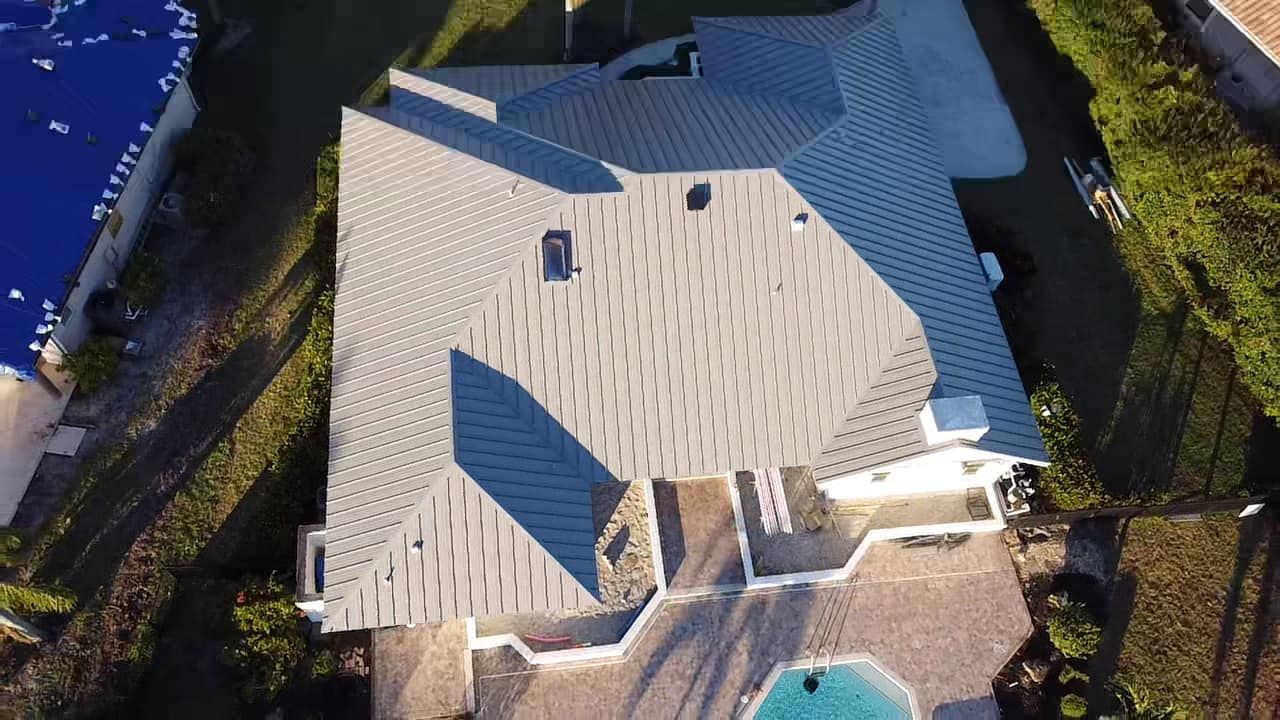 An aerial view of a gray-shingled residential roof with multiple peaks, a chimney, and a swimming pool in the yard.