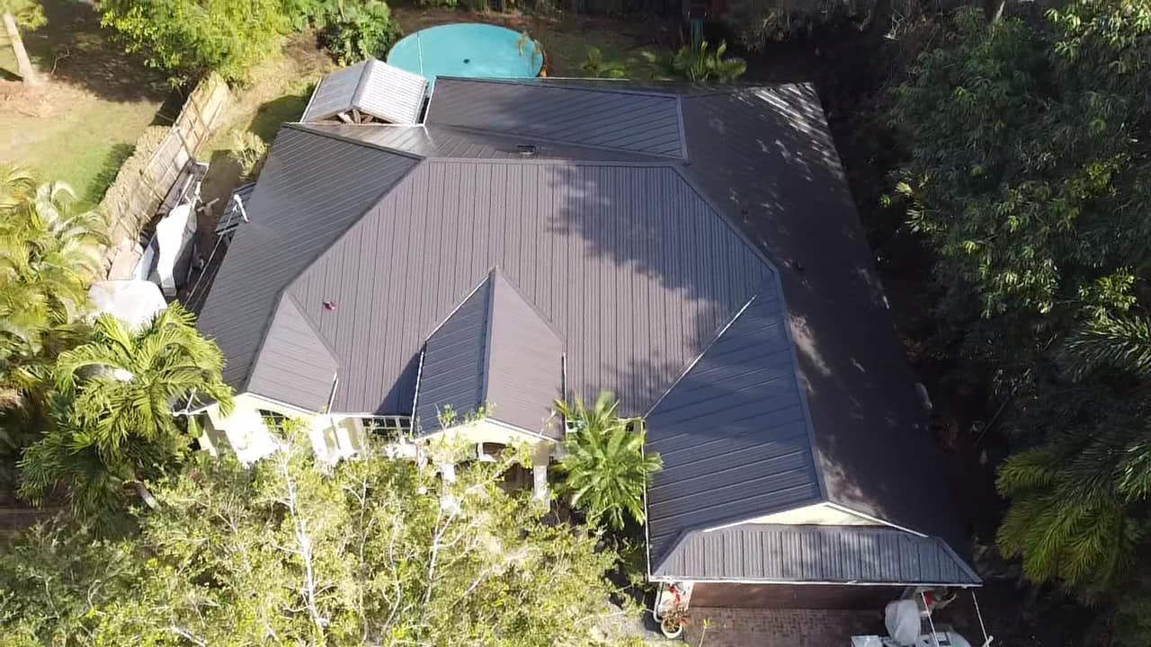 An aerial view of a dark metal-roofed house surrounded by dense green trees and a small backyard pool.