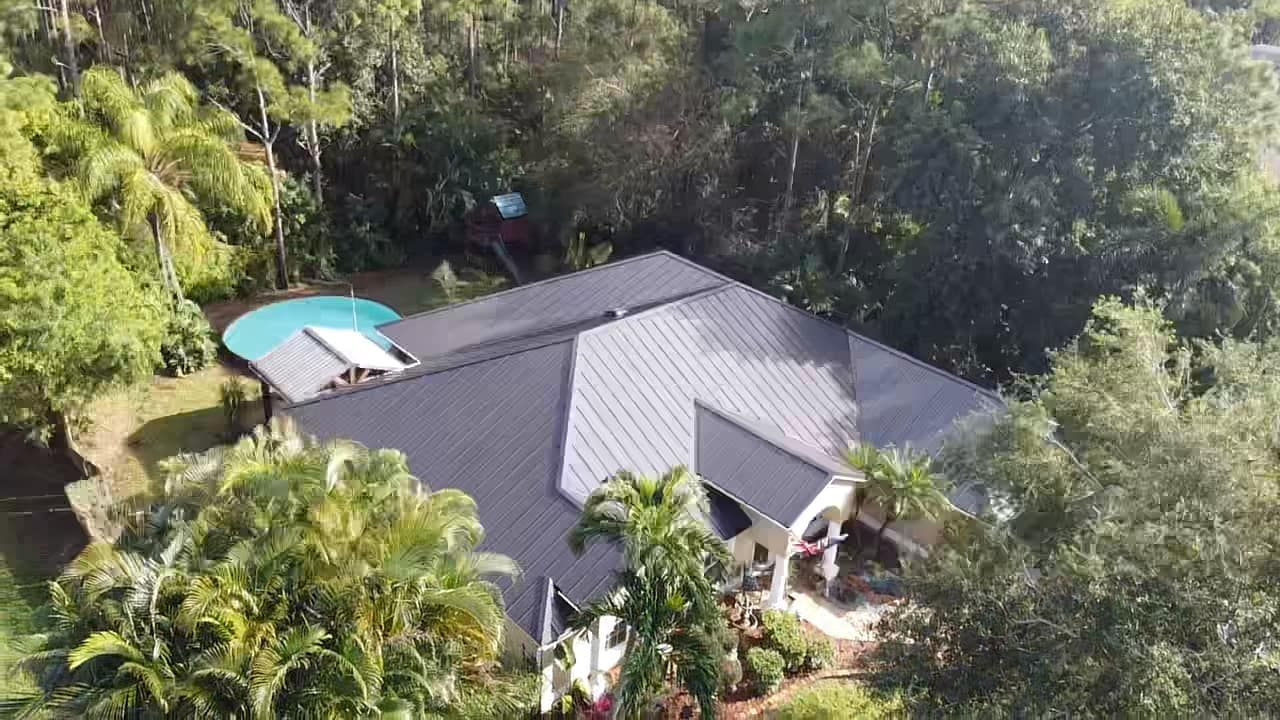 An aerial view of a suburban home with a grey roof, surrounding lush green trees, and a small pool in the backyard.