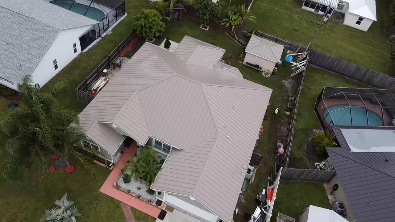 An aerial view of a single-story house with a light-colored metal roof, surrounded by lawns, fences, and swimming pools.
