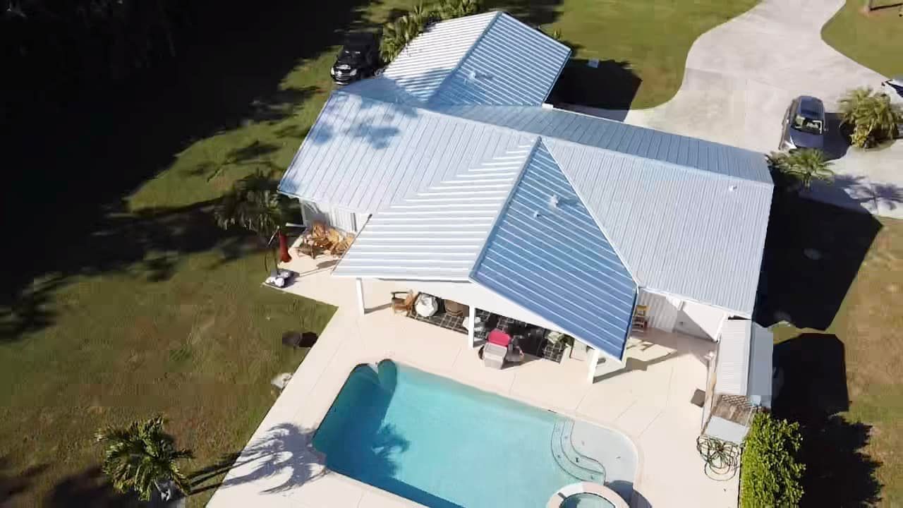 Aerial view of a white single-story home with a light blue metal roof, featuring a backyard pool and patio.