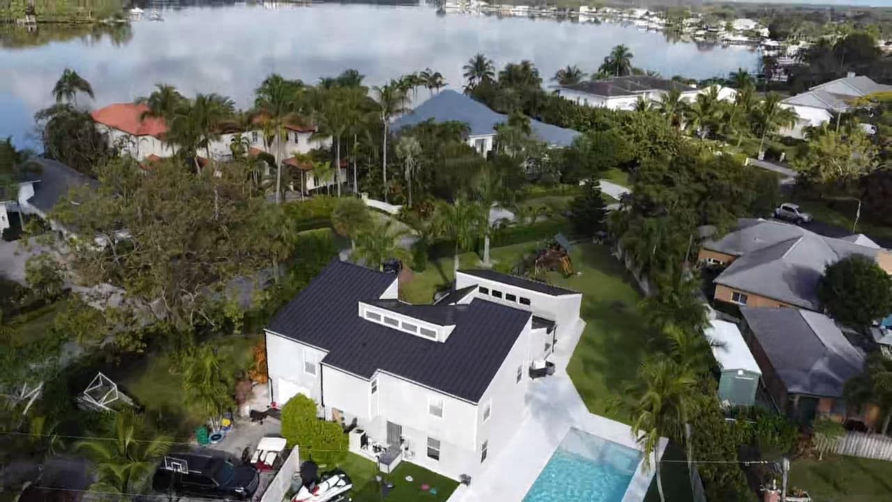 Aerial view of a white house with a dark roof, a swimming pool, and a large yard, situated near a lake in a neighborhood.