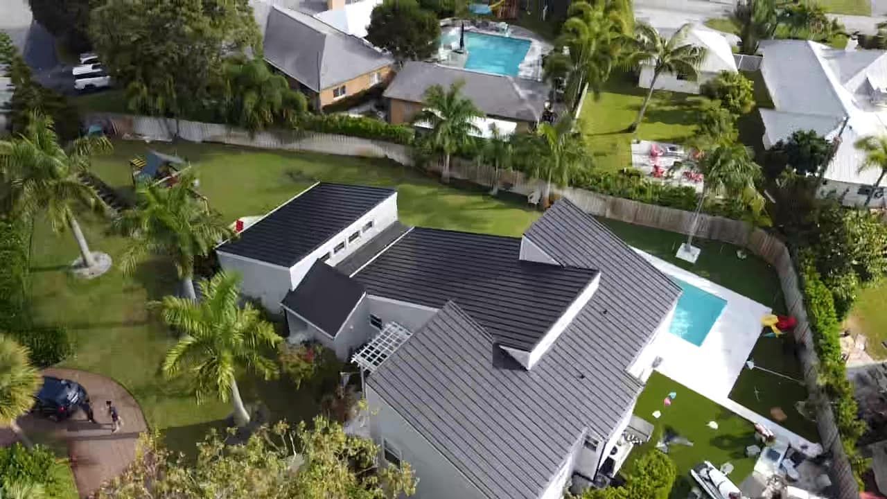 An aerial view of a single-story house with a black roof, surrounding lawn, and a swimming pool in a suburban neighborhood.