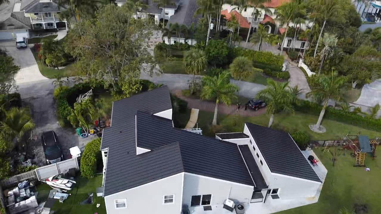 Aerial view of a white, L-shaped house with a black roof surrounded by lush green trees and a paved driveway.