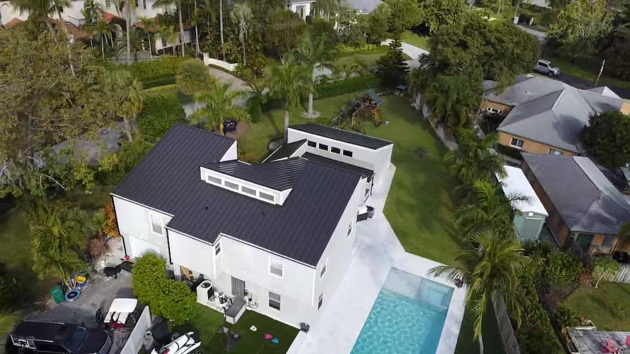 An aerial view of a white two-story house with a black metal roof, a backyard pool, and surrounding green landscaping.